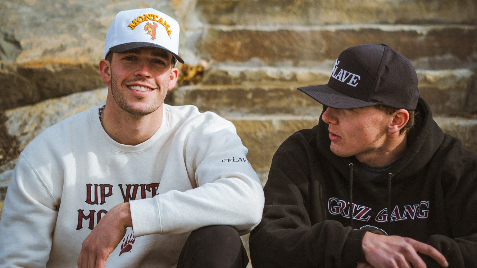 Two individuals sitting on stone steps wearing Flave branded clothing and caps.