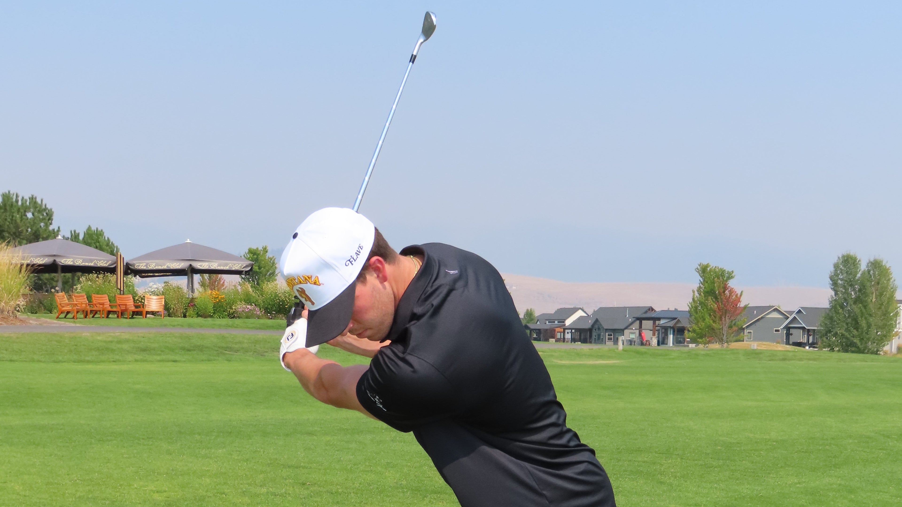 A man in a black polo shirt, black shorts, white Vintage Montana Flave cap, and white golf shoes is captured mid-swing on a golf course.