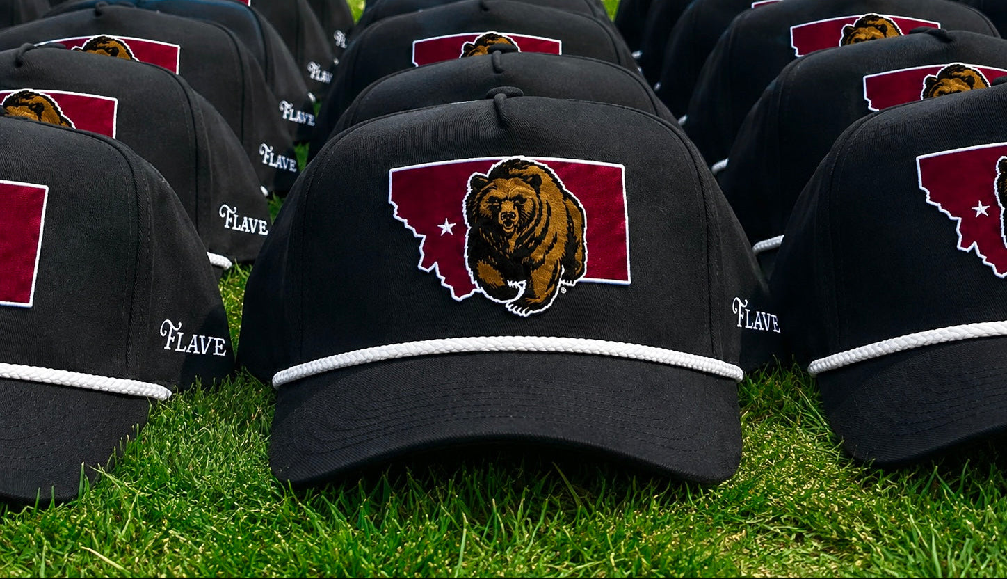 A row of black baseball caps with a maroon and white embroidered logo of a grizzly bear inside the state of Montana, displayed on a green grass field.