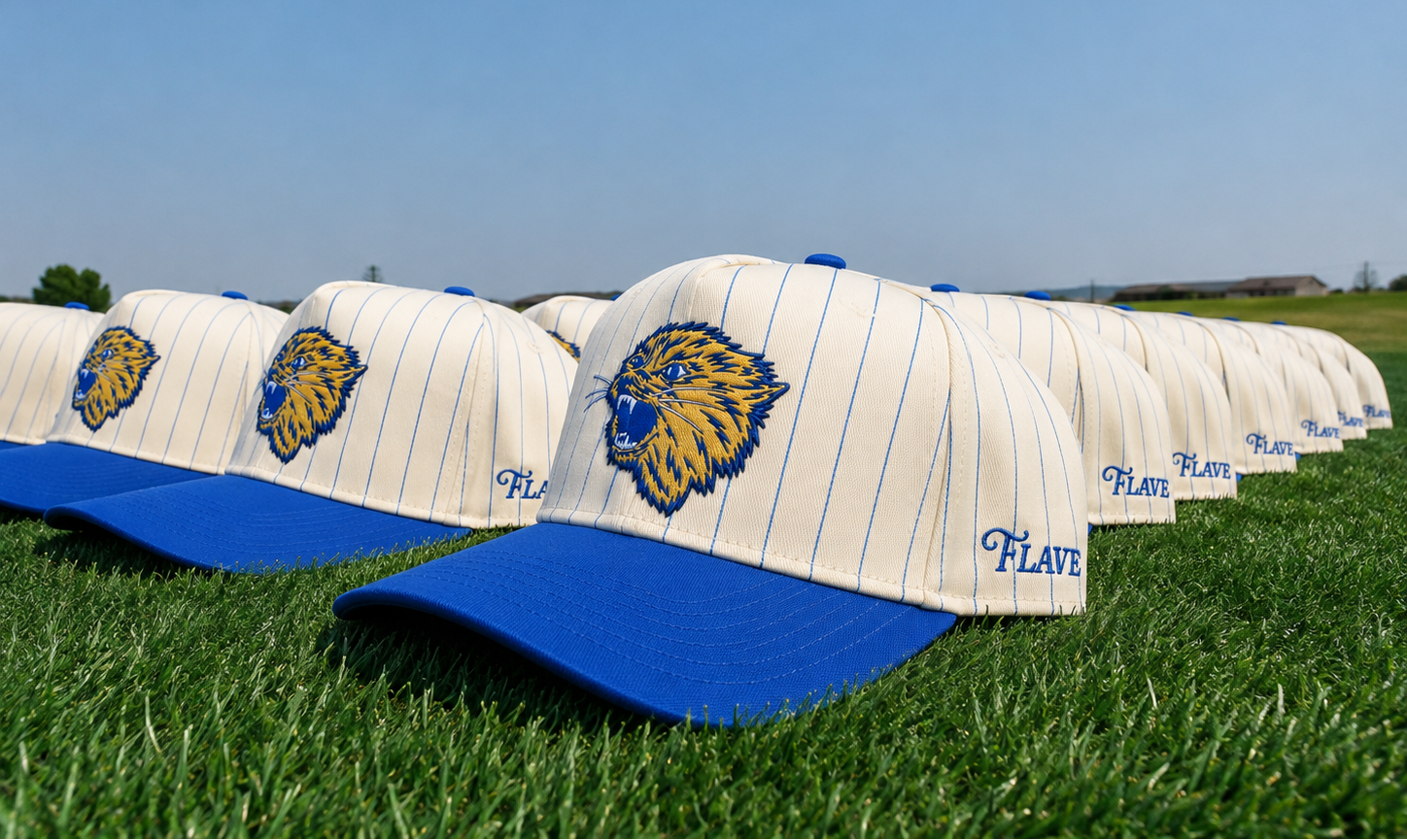A row of Montana State Vintage Cats caps with blue bills and blue pinstripes sits on green grass under a clear blue sky. Each cap features a blue and yellow Bobcat logo on the front and the name 'Flave' embroidered on the side.